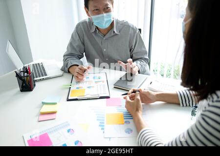 Ein Geschäftsmann, der eine Maske trägt und einem Mitarbeiter- oder Mitarbeiterteam-Diagramm auf dem Tischnotiz an der Wand i über den Geschäftsmarketingvorgang erklärt Stockfoto