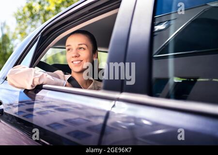 Frau im Auto als Beifahrer auf dem Rücksitz. Lächelnde Kundin im Taxi, die aus dem Fenster schaute. Glückliche elegante Geschäftsfrau. Stockfoto