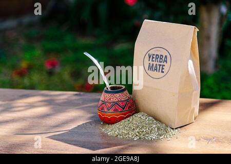 Eine rustikale Packung braunes Papier mit einem traditionellen Mate aus Kalabash über einem Holztisch mit Yerba Mate darüber verstreut. Stockfoto