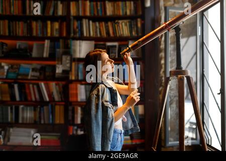 Die junge Hipster-Hündin schaut durch ein Teleskop, das im Vintage-Bibliotheksraum steht. Stockfoto