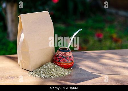 Eine rustikale Packung braunes Papier mit einem traditionellen Mate aus Kalabash über einem Holztisch mit Yerba Mate darüber verstreut. Stockfoto