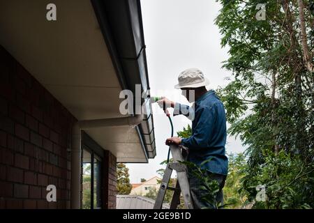 Mann, der auf der Leiter steht und die Rinne mit einem Gartenschlauch wäscht. Hauswartung. Stockfoto