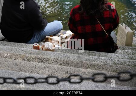 Junges Paar am Reuss-Ufer in Luzern, Schweiz. Sie essen Essen zum Mitnehmen vor einem Restaurant, das draußen sitzt. Stockfoto