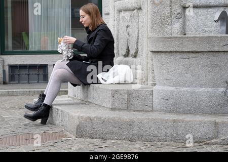 Junge Frau, die vor einem Restaurant auf einer Steintreppe Essen zum Mitnehmen isst. Sie sitzt in der Innenstadt von Luzern, Schweiz. Stockfoto