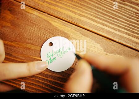 Damenhänden mit Stift schreiben die Aufschrift „Happy New Year“ auf das Etikett von Gift. Weihnachtskalligraphie. Holztisch im Hintergrund. Festliche Atmosphäre. Winterstimmung. Stockfoto