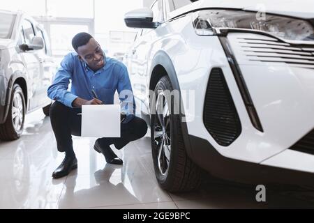 Lächelnder schwarzer Autoverkäufer, der in der Nähe des neuen Autos sitzt und in der Zwischenablage bei einem zeitgenössischen Autohändler schreibt Stockfoto