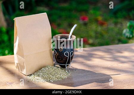 Eine rustikale Packung braunes Papier mit einem traditionellen Mate aus Kalabash über einem Holztisch mit Yerba Mate darüber verstreut. Stockfoto