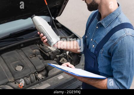 Beschnittene Ansicht des Mechanikers, der Motoröl hält und die Zwischenablage in der Nähe des Autos hält Stockfoto