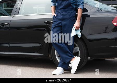 Beschnittene Ansicht eines Mechanikers, der Lappen und Hand in der Tasche mit Overalls in der Nähe des Autos im Freien hält Stockfoto