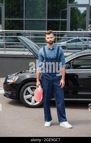 Reparaturwerkstatt in Overalls, die Scheibenwaschflüssigkeit in der Nähe des Autos im Freien halten Stockfoto