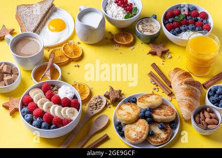 Gesunde Frühstückstisch-Szene mit Obst, Joghurts, Croissants, Smoothie, nahrhaftem Toast und Eierpfanne. Draufsicht auf gelbem Hintergrund. Stockfoto