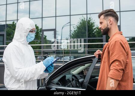 Fahrer, der in der Nähe des Autos steht und in einem Gefahrgutanzug in der Nähe eines Arzter auf der Zwischenablage steht Stockfoto