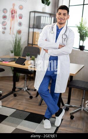 In voller Länge Porträt des jungen arabischen indischen gut aussehenden Arzt, trägt blaue Peelings und weißes Kleid stehen mit Armen gekreuzt in modernen Krankenhauszimmer. Stockfoto