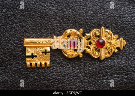 Goldener Schlüssel mit Rubinen, isoliert auf schwarzer Lederstruktur Stockfoto