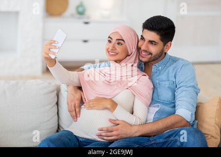 Liebevolles junges, schwanger-arabisches Paar, das Selfie auf dem Smartphone macht, während es sich zu Hause auf dem Sofa entspannt, freien Platz Stockfoto