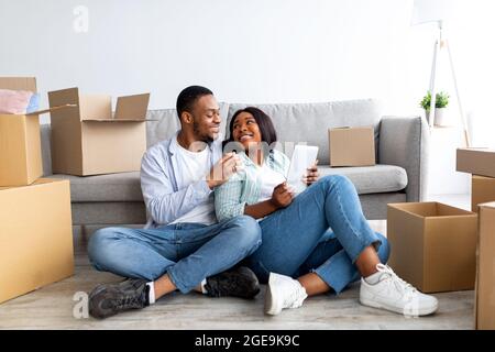 Liebevolle schwarze Familie mit digitalem Tablet und Umarmung, während sie auf dem Boden im neuen Haus unter ausgepackten Boxen sitzt Stockfoto