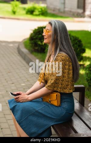 Frau mittleren Alters, grauhaarige Frau in trendigen Kleidern sitzt auf der Stadtbank mit Smartphone Stockfoto