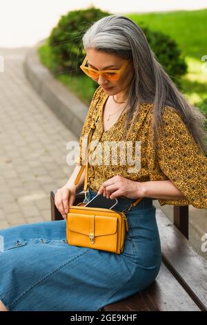 Trendige reife Frau mit Umhängetasche und Smartphone auf der Bank in der Stadt sitzen Stockfoto