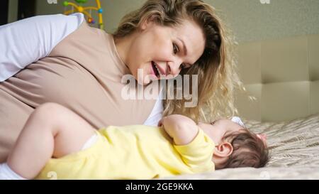 Fröhliche Mutter mit lockigen Haaren spielt mit dem Baby im Bett Stockfoto