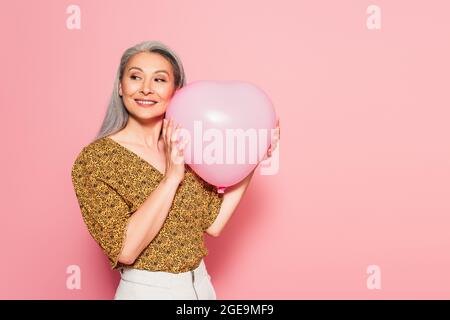 Glückliche Frau mittleren Alters in gemusterter Bluse mit herzförmigen Ballon auf rosa Hintergrund Stockfoto