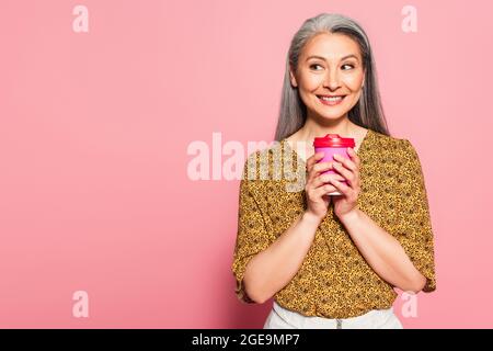 Fröhliche Frau mittleren Alters mit Kaffee, um auf rosa Hintergrund zur Seite zu schauen Stockfoto