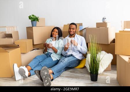 Glückliche afroamerikanische Ehegatten sitzen zwischen ausgepackten Kartons und trinken Kaffee und lächeln vor der Kamera Stockfoto