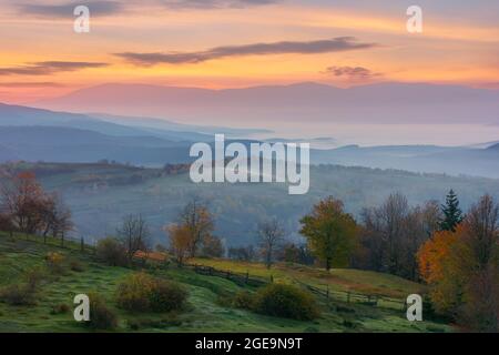 Herbstliche ländliche Landschaft. Wunderschöne Naturlandschaft mit nebligen Tal und glühenden Himmel bei Sonnenaufgang. Bäume in bunten Laub und Felder auf Hügeln am Morgen Stockfoto