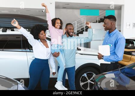 Schwarze Familie fühlt sich aufgeregt über den Kauf von Auto, Heben der Hände, gestikuling JA, stehen mit Auto Verkäufer bei Händler Stockfoto