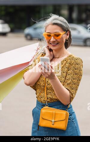 Trendige Frau mittleren Alters mit Einkaufstaschen, die auf das Handy auf der Straße schauen Stockfoto