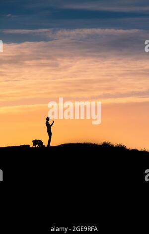 Ein Mann mit seinem Hund, der steht und mit seinem Mobiltelefon die Aussicht vom Gipfel des Pentire Point East fotografiert und von einem intensiven Sonnenuntergang in Newquay in Cornwall umrackt wird. Stockfoto