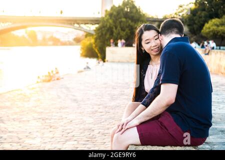 Junges multirassisches Paar sitzt am Fluss mit einem Sonnenuntergang im Hintergrund. Asiatische Freundin und europäischen Freund genießen ihre Liebe. Junge Stockfoto