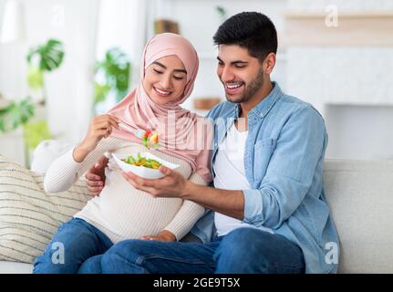 Ausgewogene Ernährung in der Schwangerschaft Konzept. Fürsorglicher muslimischer Mann, der seiner erwartungsvollen Frau leckeren Gemüsesalat gab und auf dem Sofa saß Stockfoto