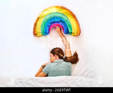 Draufsicht auf junge Frauen, die einen bunten Regenbogen-Luftballon halten, während sie unter einer Decke im Bett mit weißen Laken schlafen Stockfoto