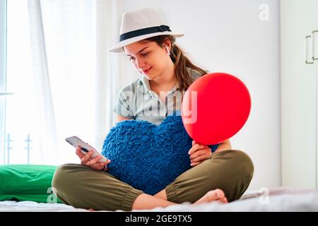 Junge Frau in legerer Kleidung und Hut mit rotem Ballon und herzförmigem Kissen beim Stöbern auf dem Smartphone, während sie allein auf dem Bett Geburtstag feiern Stockfoto