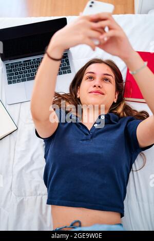 Von oben eine positive junge Studentin, die auf dem Bett liegt und Selfie auf dem Smartphone nimmt, während sie während eines Remote-Online-Studiums zu Hause Pause hat Stockfoto