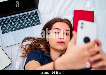 Von oben eine positive junge Studentin, die auf dem Bett liegt und Selfie auf dem Smartphone nimmt, während sie während eines Remote-Online-Studiums zu Hause Pause hat Stockfoto