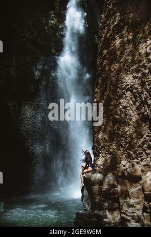 Seitenansicht der unkenntlichen weiblichen Reisenden, die sich entspannen und die frische Luft genießen, während sie am sonnigen Tag i auf einem rauen felsigen Hang in der Nähe eines plätschernden Wasserfalls sitzen Stockfoto