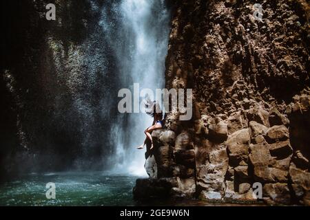 Seitenansicht der unkenntlichen weiblichen Reisenden, die sich entspannen und die frische Luft genießen, während sie am sonnigen Tag i auf einem rauen felsigen Hang in der Nähe eines plätschernden Wasserfalls sitzen Stockfoto