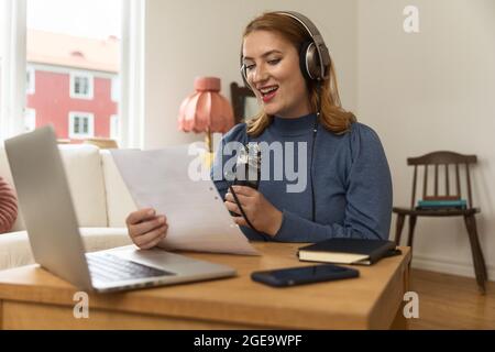 Begeisterte Radiomoderatorin, die Podcast aufzeichnete, während sie zu Hause mit dem Mikrofon und beim Lesen von Papiernotizen zugegen war Stockfoto