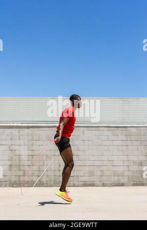 Seitenansicht des energiegeladenen afroamerikanischen männlichen Athleten, der im Sommer beim Training Seil über den Boden hüpft Stockfoto