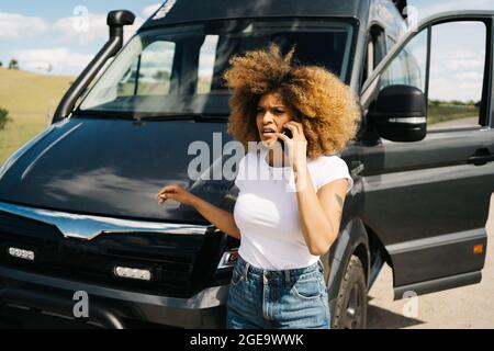 Besorgt junge afroamerikanische Frau mit lockigen Haaren reden auf dem Handy, während sie um Hilfe von Reparatur-Service nach Unfall mit Wohnmobil fragen Stockfoto