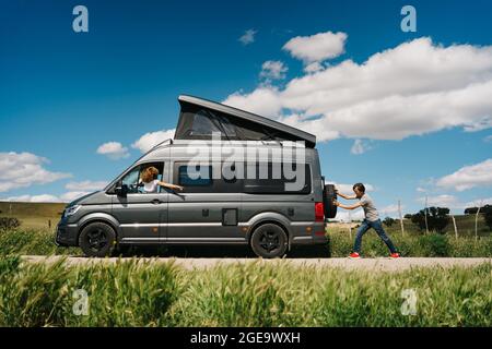 Seitenansicht des jungen Mannes, der den Wohnmobil schiebt, während die Frau am Lenkrad sitzt, weil sie während der Reise in die Sommerlandschaft Probleme hatte Stockfoto