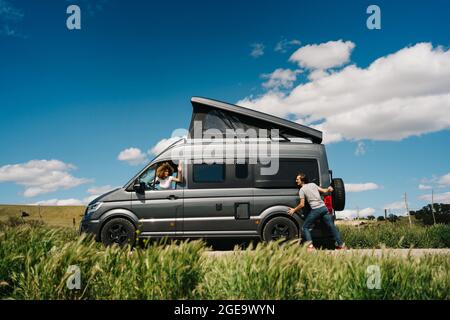 Seitenansicht des jungen Mannes, der den Wohnmobil schiebt, während die Frau am Lenkrad sitzt, weil sie während der Reise in die Sommerlandschaft Probleme hatte Stockfoto