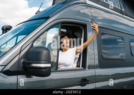 Unglückliche junge afroamerikanische Reisende, die auf dem Fahrersitz im Wohnmobil sitzt und versucht, eine Verbindung über das Mobiltelefon zu bekommen, während sie Probleme hat Stockfoto