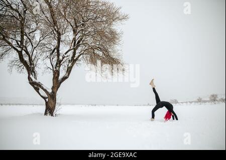 Seitenansicht einer flexiblen Frau, die in Eka Pada Urdhva Dhanurasana Yoga praktiziert, während sie im Winter mit erhobenem Bein auf einem schneebedeckten Feld steht Stockfoto