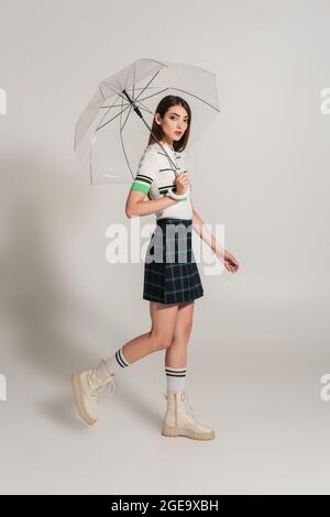 Junge Frau in stilvoller Kleidung unter transparentem Regenschirm auf grauem Hintergrund Stockfoto