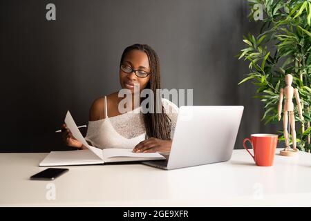 Inhalt die schwarze freiberufliche Mitarbeiterin liest Notizen in einem Notizbuch, während sie mit einem Laptop am Tisch sitzt und an einem Geschäftsprojekt im modernen Heimbüro arbeitet Stockfoto