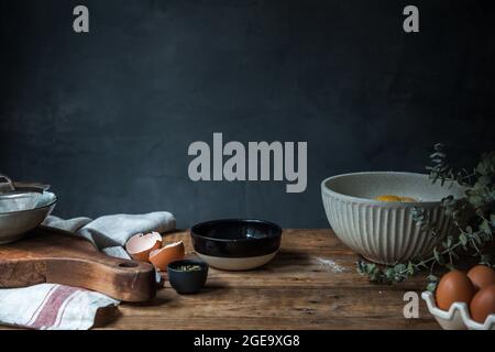 Schüssel mit Eiern, Mehl und Holzlöffel mit Kürbispüree auf Holztisch bei der Konditorei auf dunklem Hintergrund Stockfoto