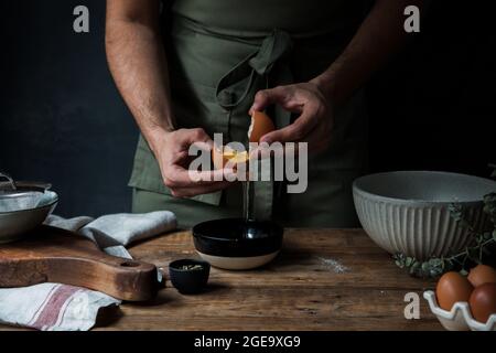 Ein nicht erkennbarer Typ in der Schürze zerbricht rohes Ei über der Schüssel, während er Gebäck auf dem Holztisch in der Nähe von Küchenutensilien zubereitete Stockfoto