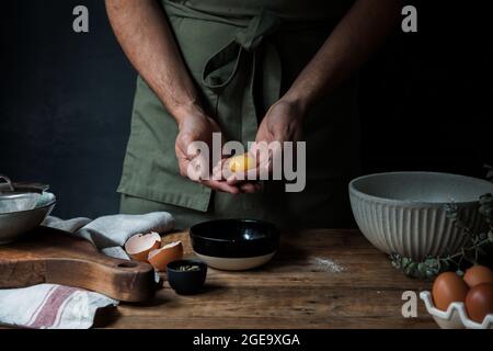 Ein nicht erkennbarer Typ in der Schürze zerbricht rohes Ei über der Schüssel, während er Gebäck auf dem Holztisch in der Nähe von Küchenutensilien zubereitete Stockfoto